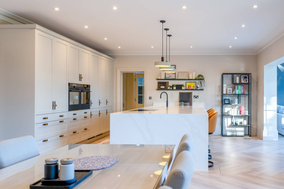 Inspiration for a large transitional single-wall open plan kitchen in Hertfordshire with a single-bowl sink, recessed-panel cabinets, grey cabinets, quartzite benchtops, black appliances, light hardwood floors, with island, brown floor and white benchtop.