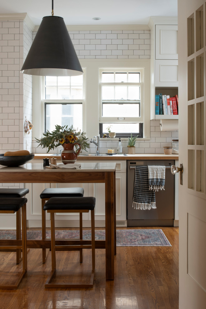 Example of a transitional galley medium tone wood floor and brown floor enclosed kitchen design in Minneapolis with a drop-in sink, recessed-panel cabinets, white cabinets, wood countertops, white backsplash, subway tile backsplash, stainless steel appliances, an island and beige countertops