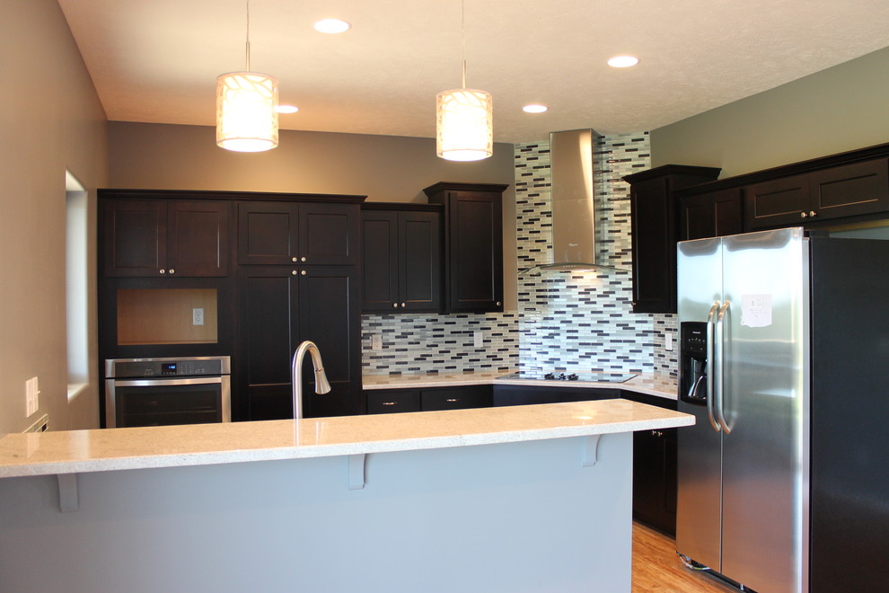 Mid-sized transitional l-shaped medium tone wood floor and brown floor kitchen photo with an undermount sink, recessed-panel cabinets, dark wood cabinets, multicolored backsplash, stainless steel appliances, a peninsula and white countertops