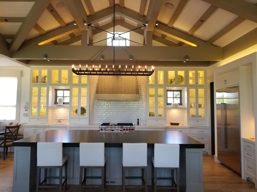 Photo of a mid-sized country single-wall eat-in kitchen in San Francisco with a farmhouse sink, glass-front cabinets, white cabinets, solid surface benchtops, white splashback, subway tile splashback, stainless steel appliances, medium hardwood floors, with island, brown floor and black benchtop.
