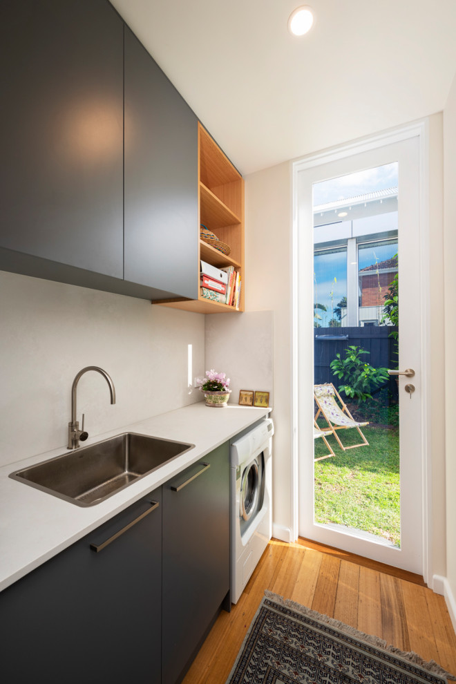 Small contemporary galley utility room in Melbourne with a drop-in sink, flat-panel cabinets, grey cabinets, granite benchtops, white splashback, granite splashback, beige walls, medium hardwood floors, an integrated washer and dryer, brown floor and white benchtop.