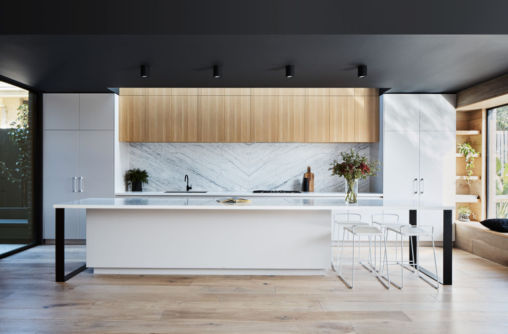 Inspiration for a contemporary galley kitchen in Melbourne with an undermount sink, flat-panel cabinets, light wood cabinets, white splashback, stone slab splashback, panelled appliances, light hardwood floors, with island, beige floor and white benchtop.