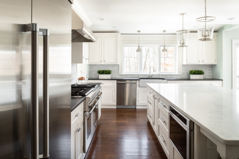 This is an example of a large transitional l-shaped kitchen in New York with a farmhouse sink, shaker cabinets, white cabinets, grey splashback, subway tile splashback, stainless steel appliances, medium hardwood floors, with island, brown floor and black benchtop.