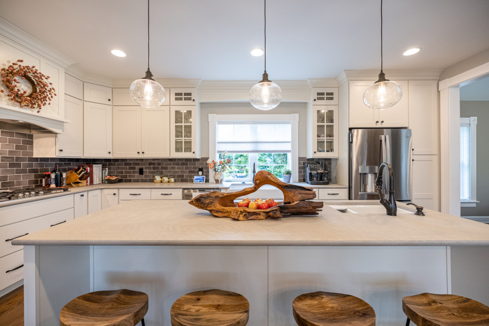 Large transitional l-shaped open plan kitchen in Cincinnati with a farmhouse sink, shaker cabinets, white cabinets, granite benchtops, grey splashback, stone tile splashback, stainless steel appliances, dark hardwood floors, with island, brown floor and grey benchtop.