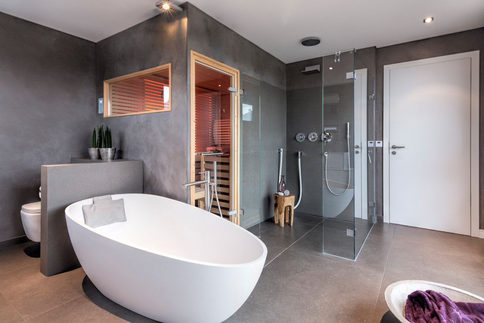 Mid-sized contemporary bathroom in Dusseldorf with flat-panel cabinets, white cabinets, a freestanding tub, a curbless shower, a wall-mount toilet, gray tile, grey walls, with a sauna, grey floor, cement tiles and a hinged shower door.
