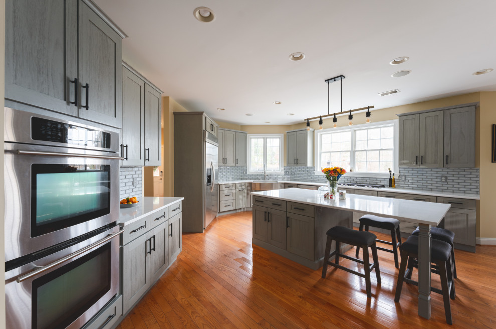Example of a large transitional u-shaped medium tone wood floor and brown floor eat-in kitchen design with a farmhouse sink, shaker cabinets, gray cabinets, quartz countertops, gray backsplash, glass tile backsplash, stainless steel appliances, an island and white countertops