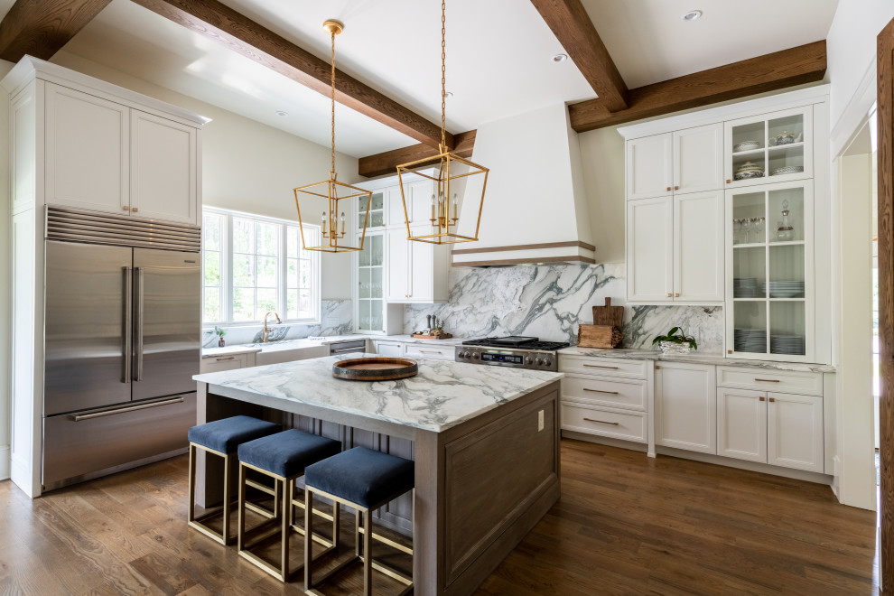 This is an example of a traditional kitchen/diner in Atlanta with a belfast sink, recessed-panel cabinets, white cabinets, marble worktops, white splashback, marble splashback, stainless steel appliances, medium hardwood flooring, an island, brown floors, white worktops and exposed beams.