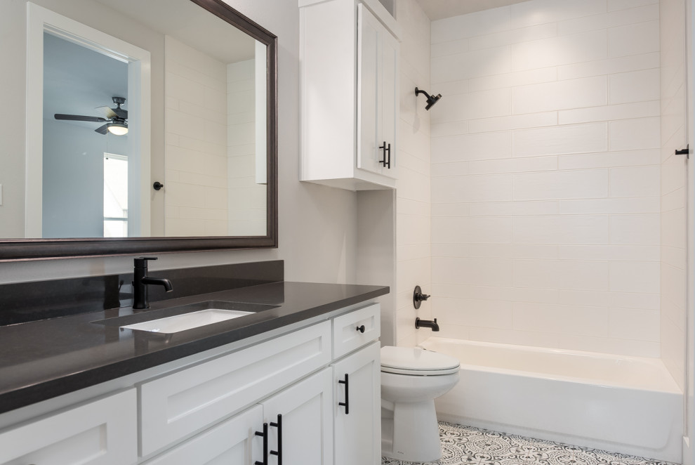 This is an example of a small country 3/4 bathroom in Dallas with raised-panel cabinets, white cabinets, a drop-in tub, a shower/bathtub combo, a one-piece toilet, white tile, subway tile, white walls, ceramic floors, an undermount sink, engineered quartz benchtops, multi-coloured floor, a shower curtain, black benchtops, a single vanity and a built-in vanity.