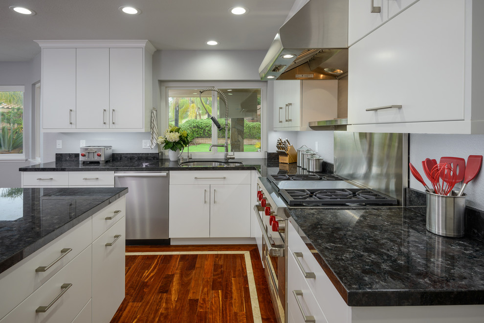 Large modern l-shaped eat-in kitchen in Los Angeles with flat-panel cabinets, white cabinets, stainless steel appliances, medium hardwood floors, with island, a drop-in sink, granite benchtops, black splashback and slate splashback.