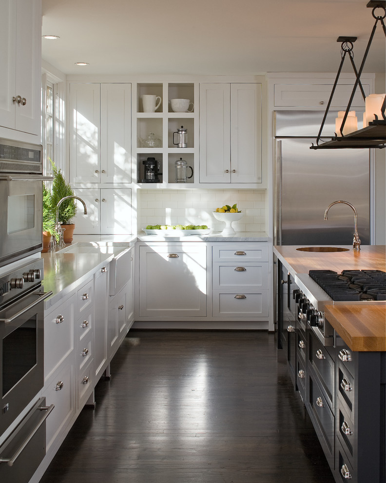 Photo of a large country l-shaped separate kitchen in San Diego with shaker cabinets, with island, a farmhouse sink, white cabinets, wood benchtops, white splashback, subway tile splashback, stainless steel appliances, dark hardwood floors and brown floor.