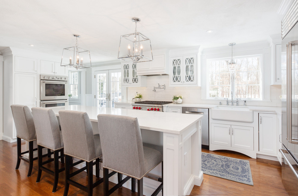 Photo of a large traditional u-shaped kitchen in Boston with white cabinets, quartz benchtops, with island, an integrated sink, shaker cabinets, white splashback, stainless steel appliances, medium hardwood floors, brown floor and white benchtop.