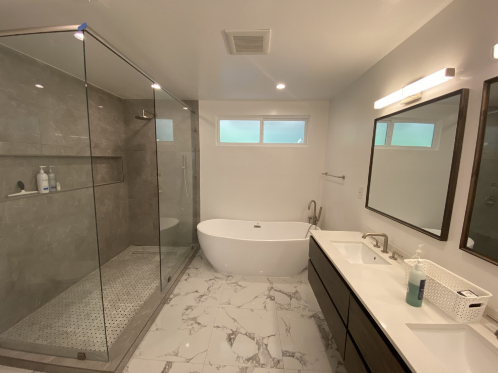 Foto på ett stort funkis vit en-suite badrum, med släta luckor, bruna skåp, ett fristående badkar, en öppen dusch, en toalettstol med hel cisternkåpa, grå kakel, keramikplattor, vita väggar, marmorgolv, ett nedsänkt handfat, marmorbänkskiva, vitt golv och med dusch som är öppen