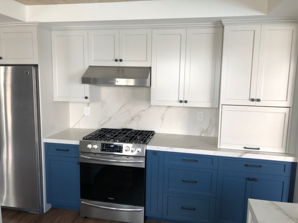 Mid-sized traditional u-shaped eat-in kitchen in Los Angeles with a farmhouse sink, shaker cabinets, blue cabinets, quartz benchtops, white splashback, engineered quartz splashback, stainless steel appliances, laminate floors, a peninsula, brown floor and white benchtop.