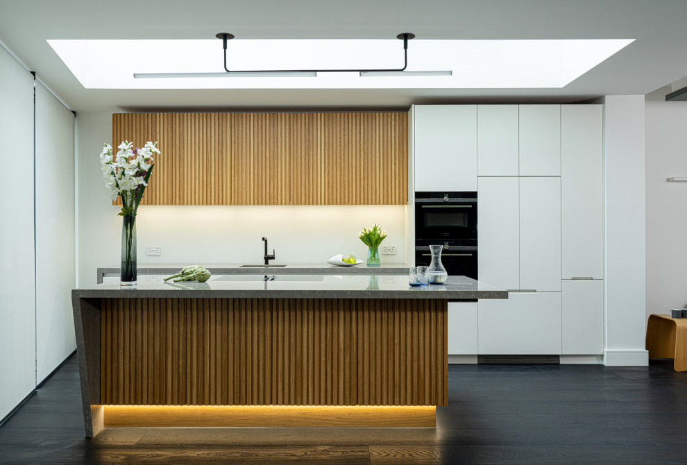 This is an example of a medium sized contemporary grey and white single-wall open plan kitchen in London with an integrated sink, flat-panel cabinets, medium wood cabinets, composite countertops, black appliances, an island, grey worktops and feature lighting.