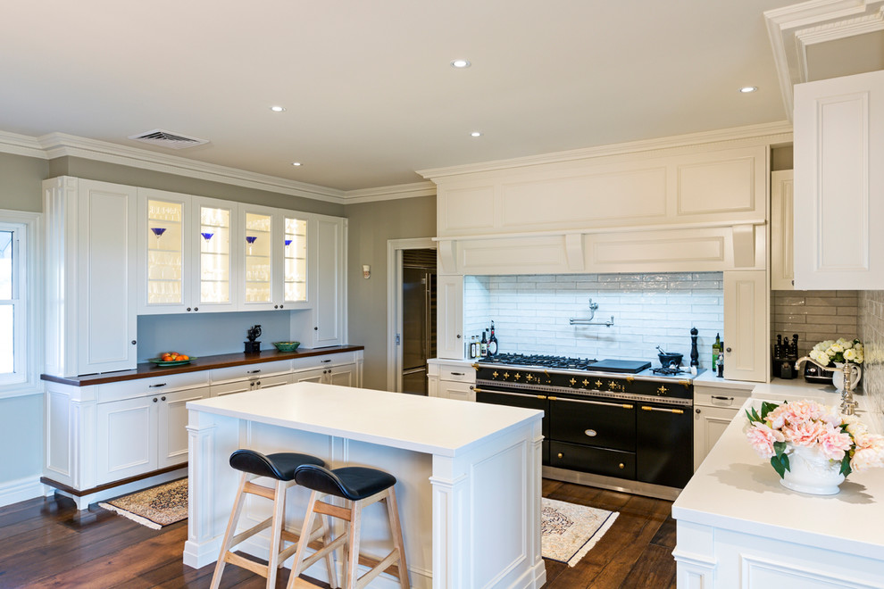 Design ideas for a transitional u-shaped eat-in kitchen in Melbourne with a farmhouse sink, recessed-panel cabinets, white cabinets, white splashback, subway tile splashback, black appliances, dark hardwood floors, with island, brown floor and white benchtop.