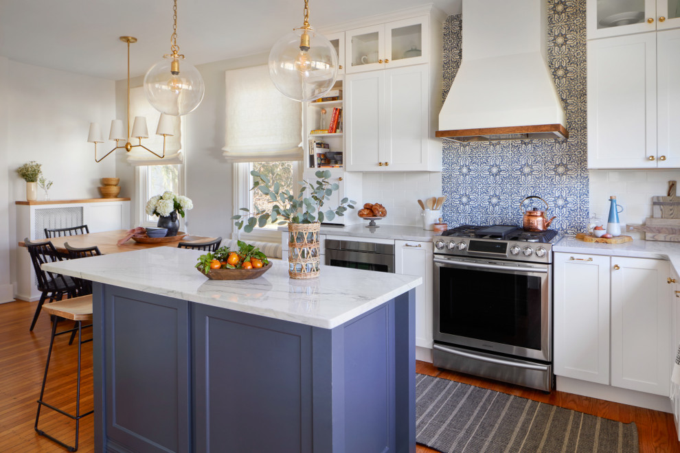 This is an example of a large country u-shaped eat-in kitchen in Philadelphia with a farmhouse sink, shaker cabinets, white cabinets, quartzite benchtops, white splashback, terra-cotta splashback, stainless steel appliances, medium hardwood floors, with island, brown floor and white benchtop.