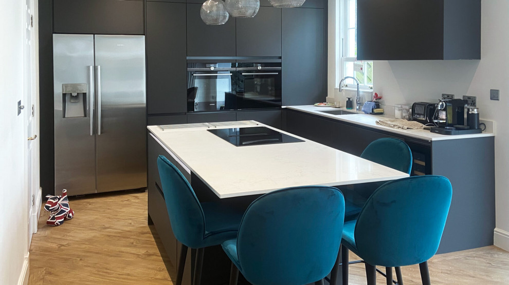Photo of a small modern l-shaped open plan kitchen in London with a single-bowl sink, flat-panel cabinets, grey cabinets, quartzite benchtops, white splashback, black appliances, light hardwood floors, with island, beige floor and white benchtop.