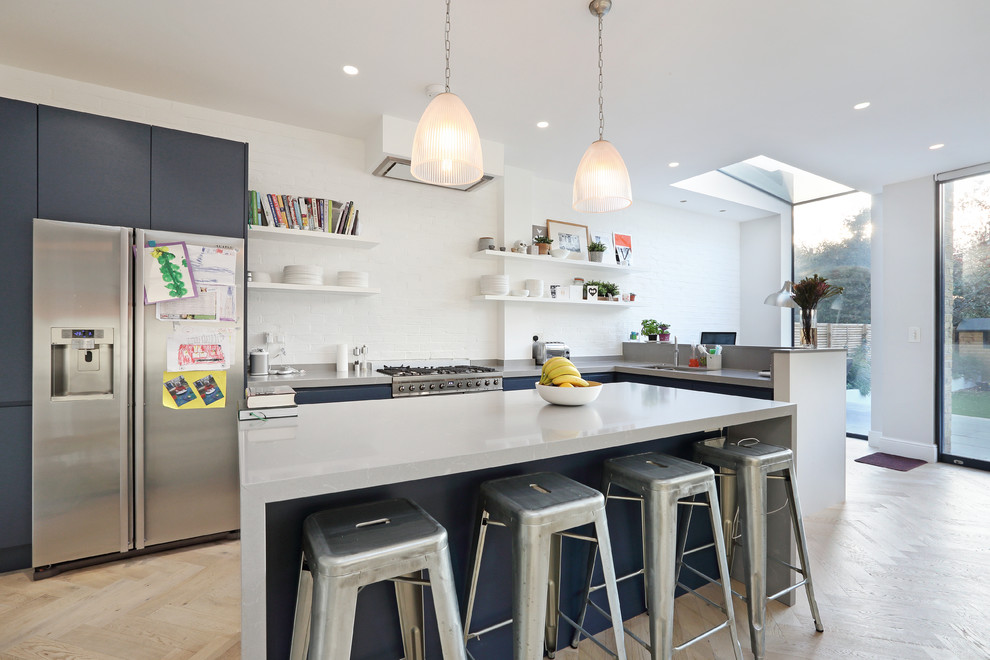 This is an example of a mid-sized contemporary l-shaped kitchen in London with flat-panel cabinets, blue cabinets, white splashback, brick splashback, stainless steel appliances, light hardwood floors, with island, beige floor, grey benchtop and an undermount sink.
