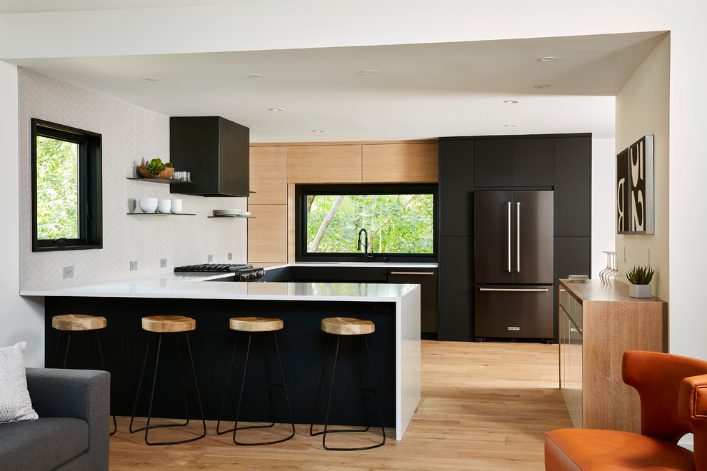 This is an example of a contemporary u-shaped open plan kitchen in Minneapolis with flat-panel cabinets, black cabinets, grey splashback, stainless steel appliances, light hardwood floors, a peninsula, beige floor and white benchtop.