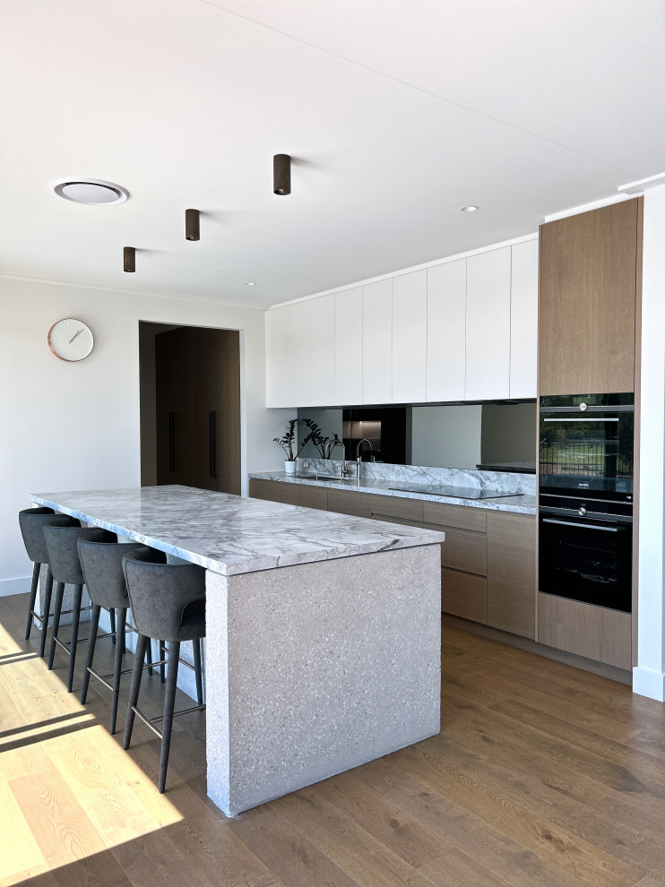 This is an example of a large modern l-shaped kitchen pantry in Sydney with flat-panel cabinets, white cabinets, marble benchtops, black splashback, mirror splashback, black appliances, with island, grey benchtop, a double-bowl sink, medium hardwood floors and brown floor.
