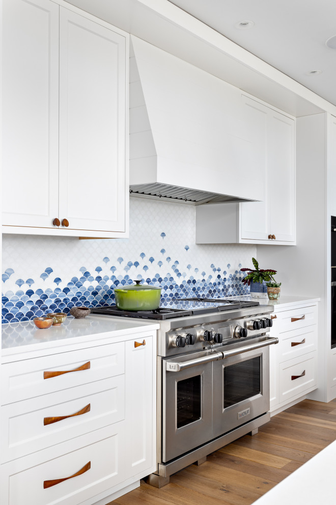 Inspiration for an expansive beach style galley open plan kitchen in Other with an undermount sink, shaker cabinets, white cabinets, quartz benchtops, blue splashback, glass tile splashback, black appliances, medium hardwood floors, with island, brown floor and white benchtop.