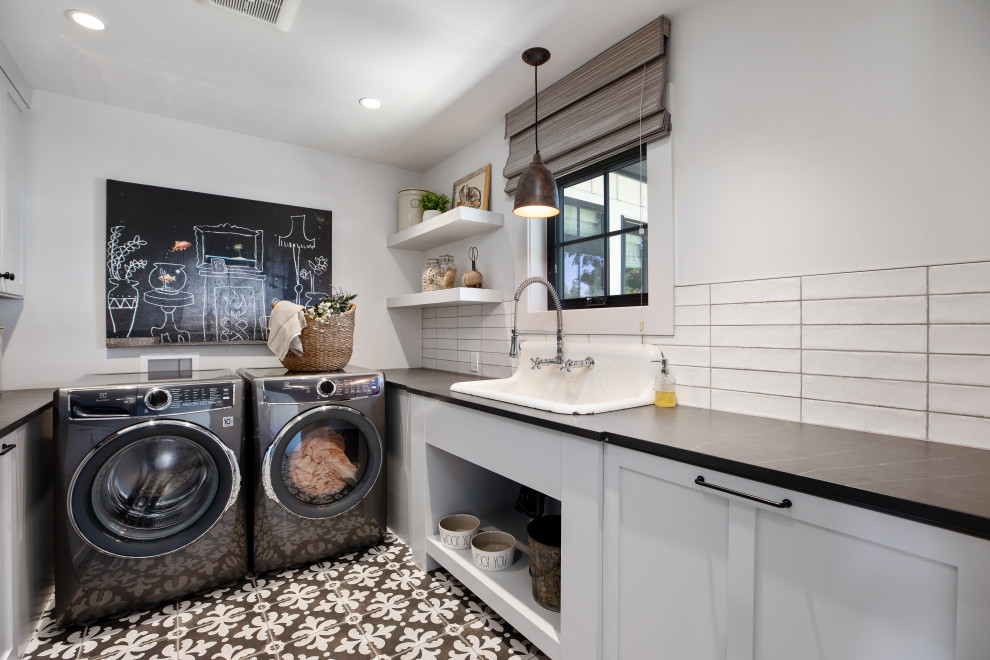 Mid-sized country single-wall dedicated laundry room in Portland with a farmhouse sink, shaker cabinets, grey cabinets, quartz benchtops, white splashback, ceramic splashback, white walls, ceramic floors, a side-by-side washer and dryer, black floor and black benchtop.