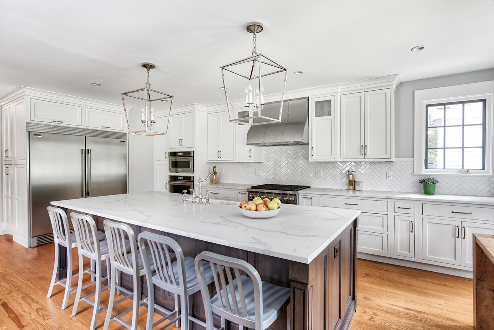 This is an example of an expansive transitional l-shaped eat-in kitchen in New York with a farmhouse sink, white cabinets, quartzite benchtops, white splashback, subway tile splashback, stainless steel appliances, medium hardwood floors, with island, white benchtop, brown floor and shaker cabinets.