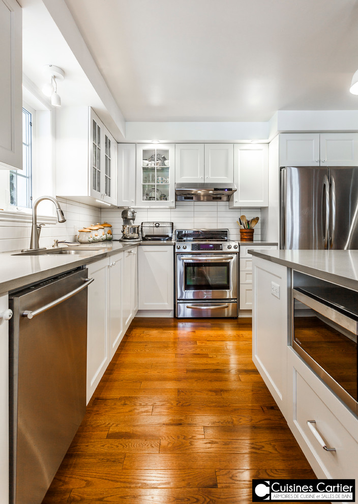 Eat-in kitchen - large transitional medium tone wood floor eat-in kitchen idea in Montreal with a double-bowl sink, shaker cabinets, white cabinets, quartzite countertops, white backsplash, ceramic backsplash, stainless steel appliances and an island
