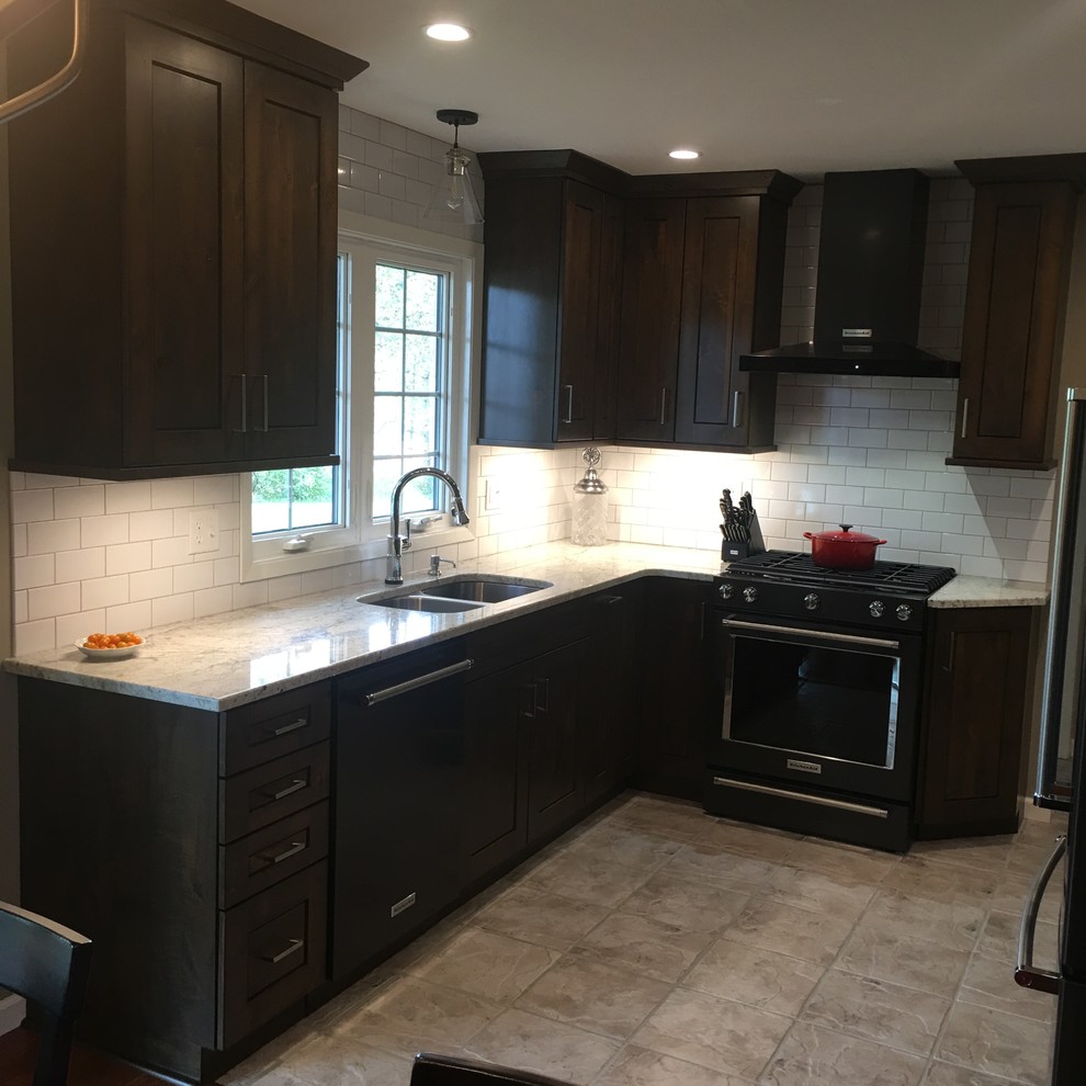 Large transitional l-shaped travertine floor and beige floor enclosed kitchen photo with a double-bowl sink, shaker cabinets, dark wood cabinets, marble countertops, white backsplash, subway tile backsplash, black appliances and an island