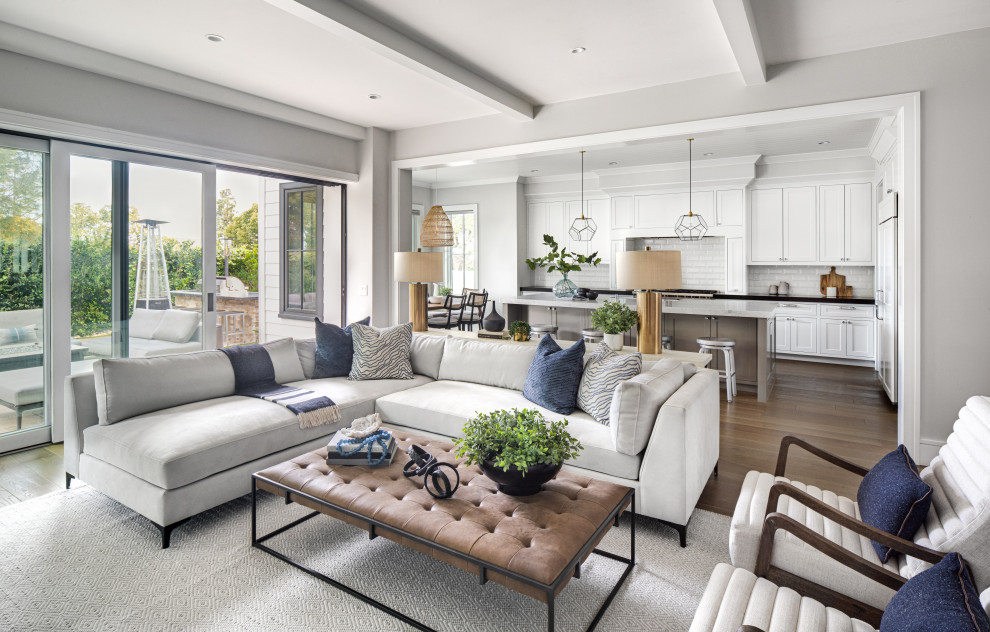 Large transitional open concept living room in Orange County with grey walls, light hardwood floors, brown floor, a standard fireplace, a wood fireplace surround, a wall-mounted tv and exposed beam.
