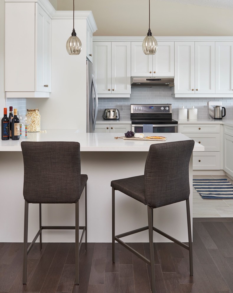 Eat-in kitchen - mid-sized transitional dark wood floor and brown floor eat-in kitchen idea in Toronto with white cabinets, solid surface countertops, glass tile backsplash, stainless steel appliances, white countertops, shaker cabinets, gray backsplash and a peninsula