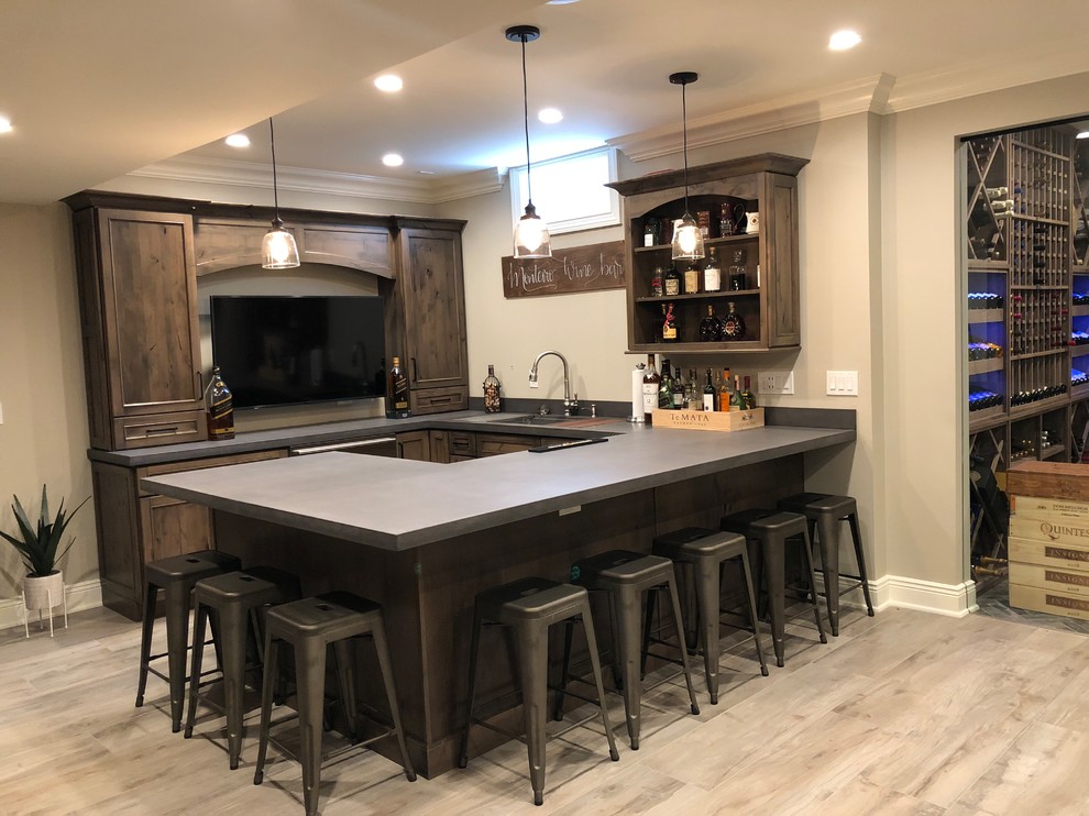 This is an example of a mid-sized country u-shaped seated home bar in New York with an undermount sink, recessed-panel cabinets, dark wood cabinets, solid surface benchtops, light hardwood floors, beige floor and grey benchtop.
