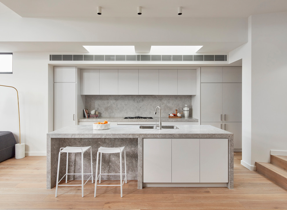 This is an example of a contemporary galley open plan kitchen in Melbourne with a double-bowl sink, flat-panel cabinets, white cabinets, medium hardwood floors, multiple islands, brown floor, marble benchtops, grey splashback, marble splashback and grey benchtop.