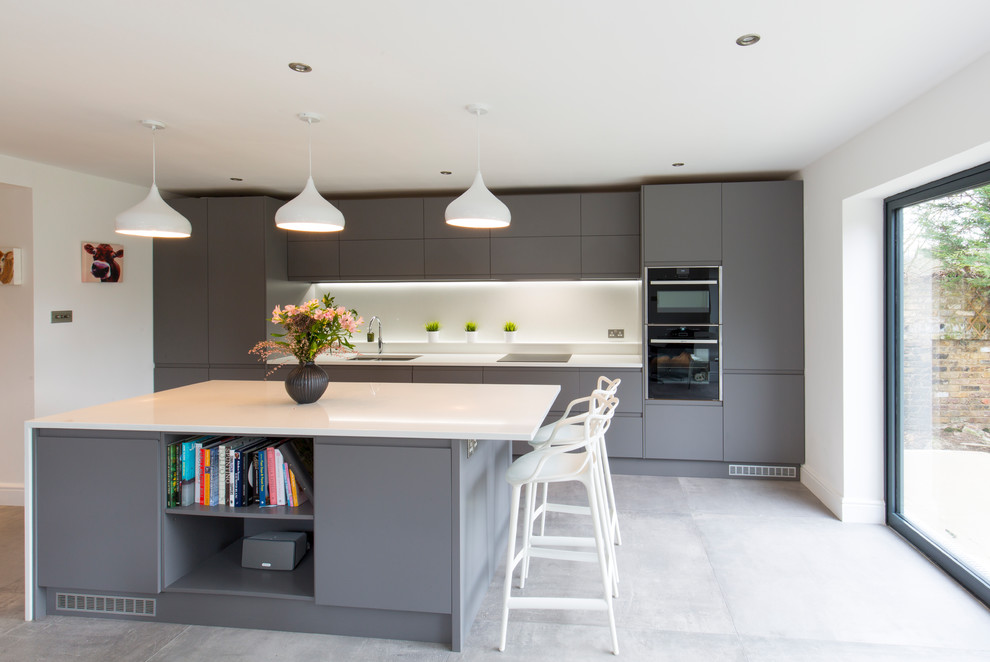 Design ideas for a large contemporary single-wall kitchen in London with flat-panel cabinets, grey cabinets, white splashback, black appliances, with island, grey floor and white benchtop.