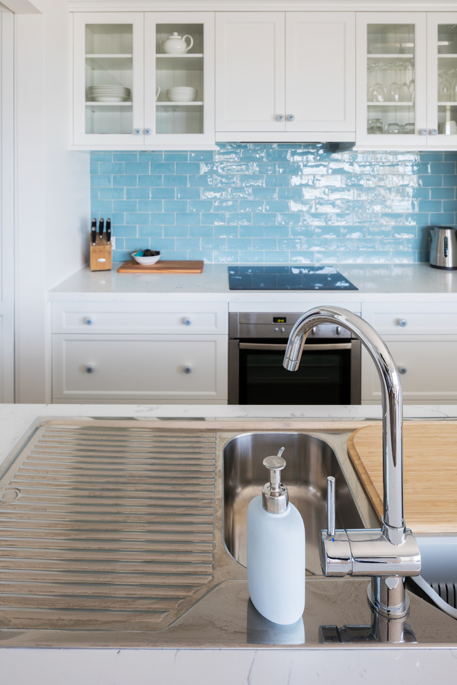 Photo of a beach style galley kitchen in Perth with a drop-in sink, shaker cabinets, white cabinets, quartz benchtops, blue splashback, ceramic splashback, stainless steel appliances, with island and white benchtop.
