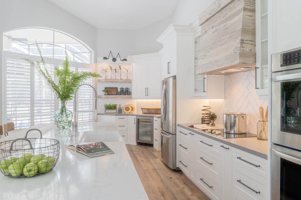 Photo of a large industrial single-wall open plan kitchen in Miami with a farmhouse sink, shaker cabinets, white cabinets, quartzite benchtops, white splashback, subway tile splashback, stainless steel appliances, vinyl floors, with island, beige floor and grey benchtop.