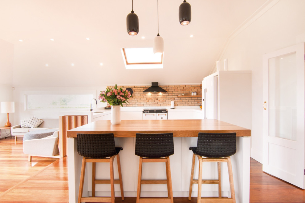 Small scandinavian u-shaped open plan kitchen in Auckland with an undermount sink, flat-panel cabinets, white cabinets, wood benchtops, red splashback, brick splashback, white appliances, with island, brown floor and brown benchtop.