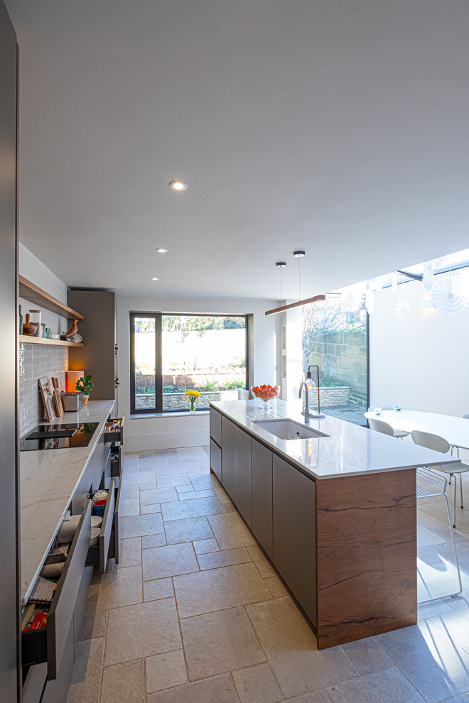 Inspiration for a medium sized contemporary galley open plan kitchen with a belfast sink, flat-panel cabinets, brown cabinets, quartz worktops, white splashback, ceramic splashback, black appliances, limestone flooring, an island, beige floors and white worktops.