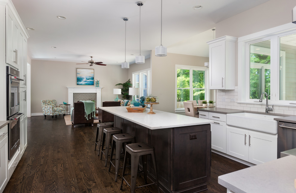 Example of a mid-sized transitional dark wood floor and brown floor open concept kitchen design in Chicago with a farmhouse sink, shaker cabinets, white cabinets, white backsplash, subway tile backsplash, stainless steel appliances, an island, gray countertops and quartz countertops