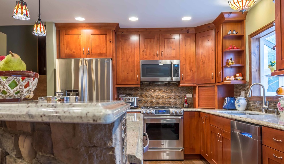 Mid-sized country l-shaped eat-in kitchen in Chicago with an undermount sink, shaker cabinets, medium wood cabinets, granite benchtops, brown splashback, glass tile splashback, stainless steel appliances, ceramic floors, with island, brown floor, grey benchtop and wallpaper.