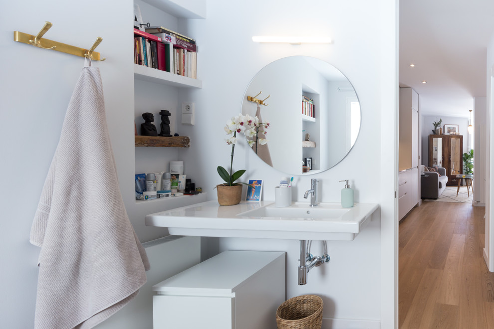 Photo of a small contemporary powder room in Madrid with flat-panel cabinets, white cabinets, white walls, medium hardwood floors, a wall-mount sink and brown floor.