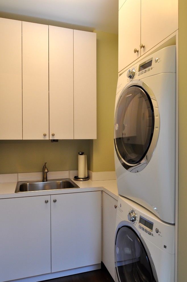 Inspiration for a mid-sized transitional l-shaped laundry cupboard in New York with a drop-in sink, flat-panel cabinets, white cabinets, green walls, dark hardwood floors and a stacked washer and dryer.