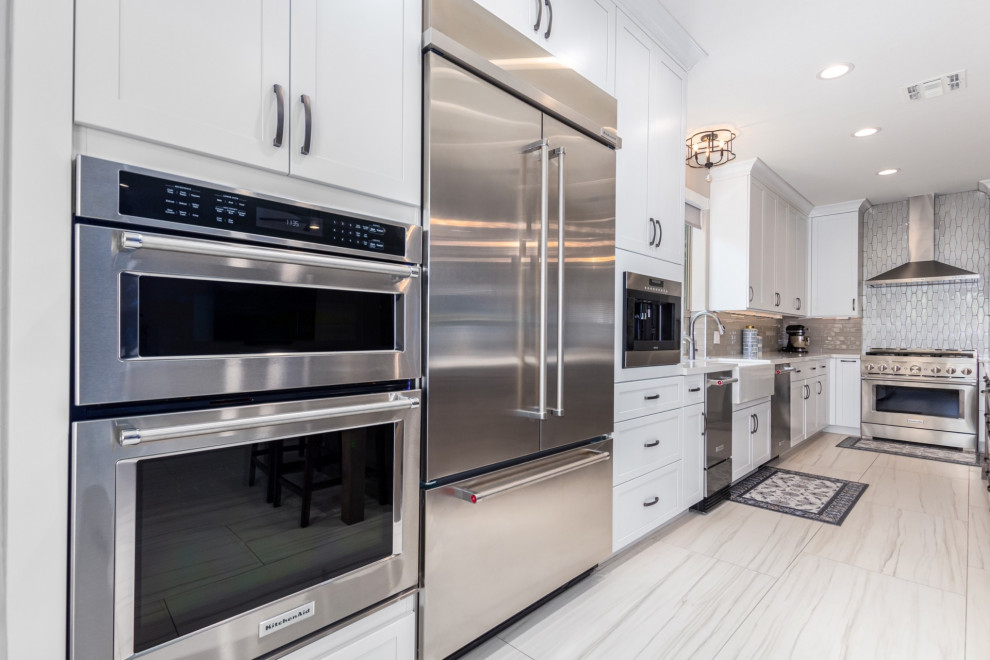 Example of a transitional kitchen design in Phoenix with a farmhouse sink, shaker cabinets, white cabinets, gray backsplash, stainless steel appliances and white countertops
