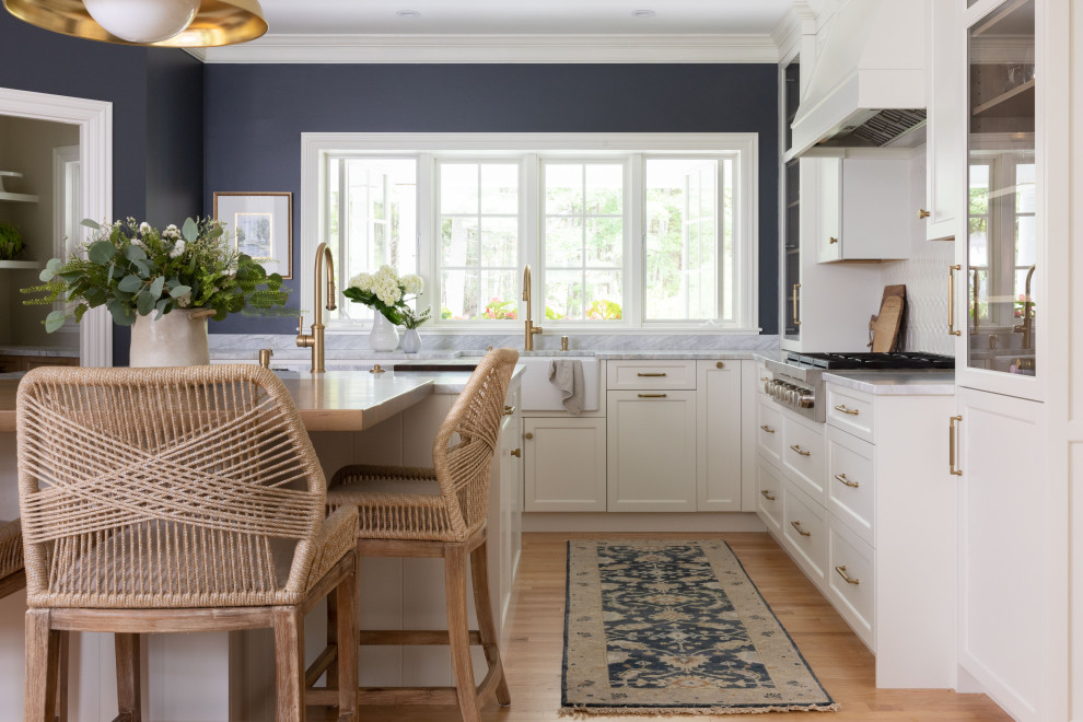 Mid-sized traditional l-shaped eat-in kitchen in Portland Maine with a farmhouse sink, flat-panel cabinets, white cabinets, marble benchtops, white splashback, ceramic splashback, stainless steel appliances, light hardwood floors, with island and blue benchtop.