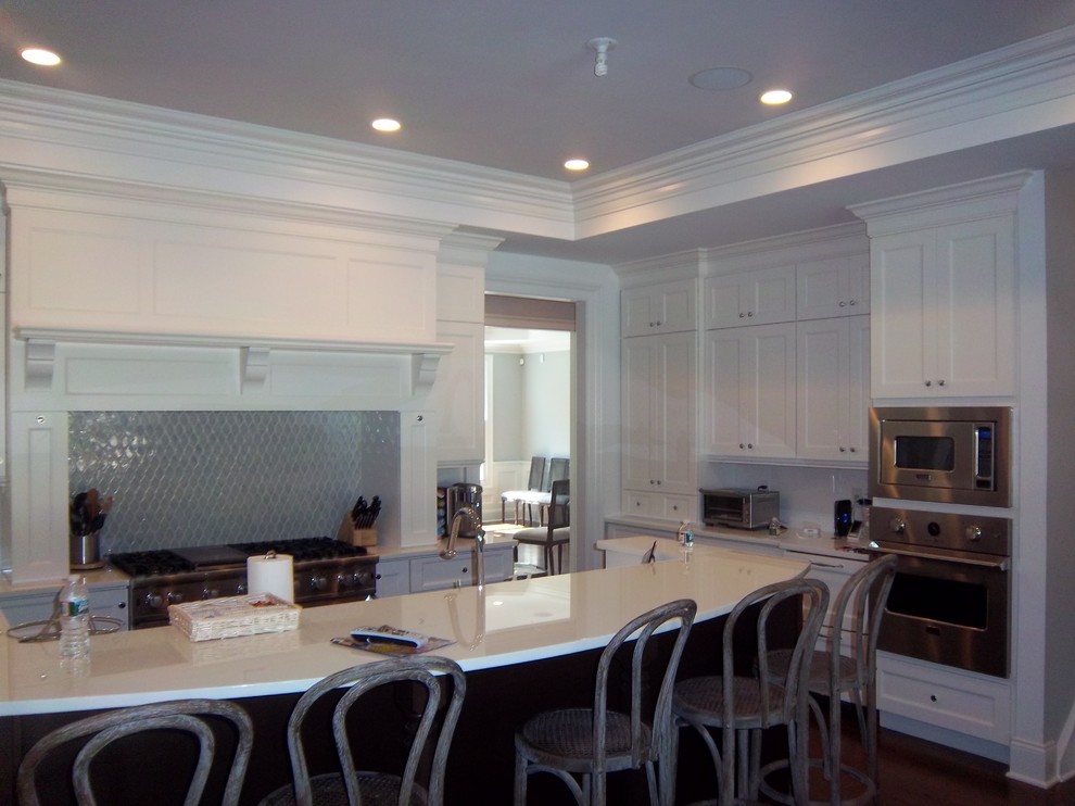 Large transitional l-shaped dark wood floor and brown floor eat-in kitchen photo with a farmhouse sink, shaker cabinets, white cabinets, solid surface countertops, white backsplash, stainless steel appliances and an island