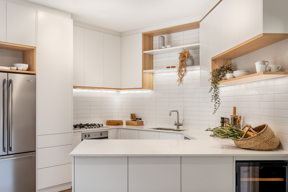 Photo of a small contemporary u-shaped eat-in kitchen in Adelaide with an undermount sink, flat-panel cabinets, white cabinets, quartz benchtops, white splashback, subway tile splashback, stainless steel appliances, laminate floors, a peninsula, brown floor and white benchtop.