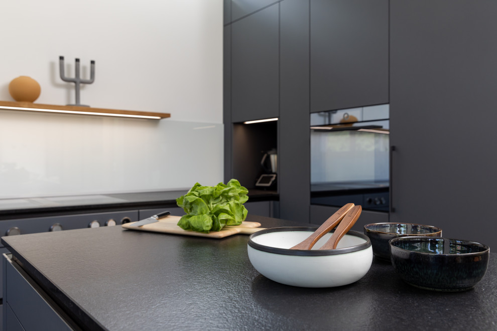 This is an example of a mid-sized contemporary u-shaped open plan kitchen in Frankfurt with an undermount sink, flat-panel cabinets, grey cabinets, granite benchtops, white splashback, glass sheet splashback, black appliances, medium hardwood floors, with island, brown floor, black benchtop and recessed.