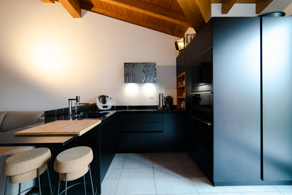 This is an example of a mid-sized modern u-shaped eat-in kitchen in Milan with a drop-in sink, flat-panel cabinets, black cabinets, quartz benchtops, grey splashback, glass sheet splashback, black appliances, porcelain floors, a peninsula, grey floor, black benchtop and exposed beam.