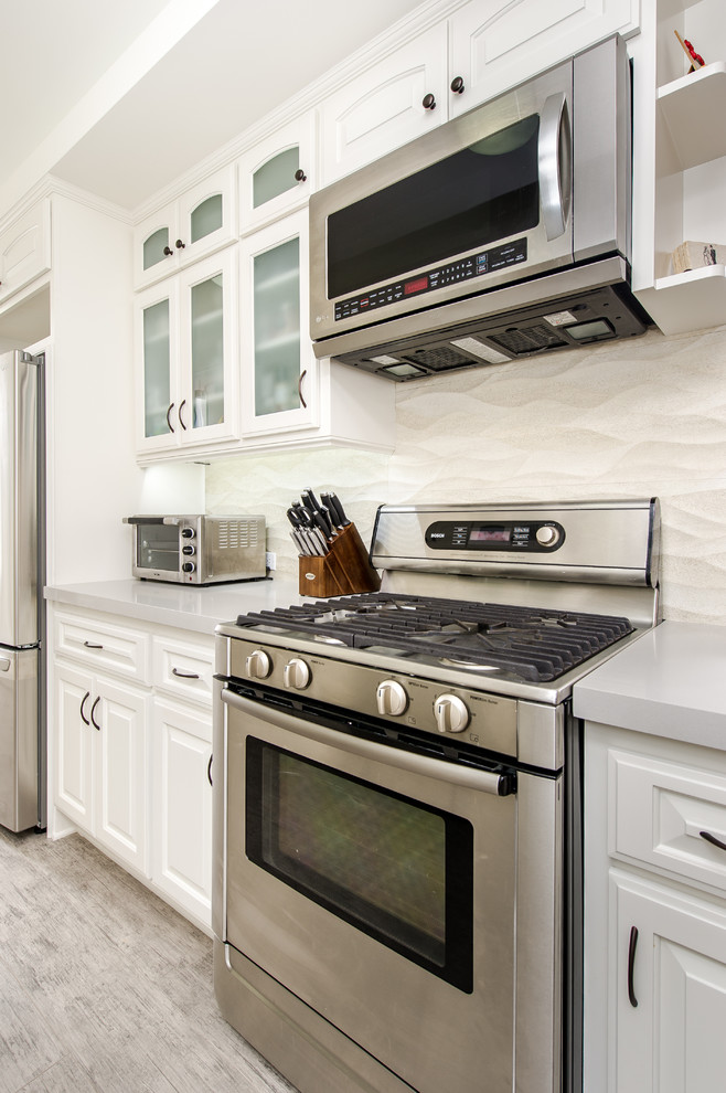 This is an example of a small traditional galley separate kitchen in Los Angeles with an undermount sink, glass-front cabinets, white cabinets, quartz benchtops, beige splashback, ceramic splashback, stainless steel appliances and porcelain floors.