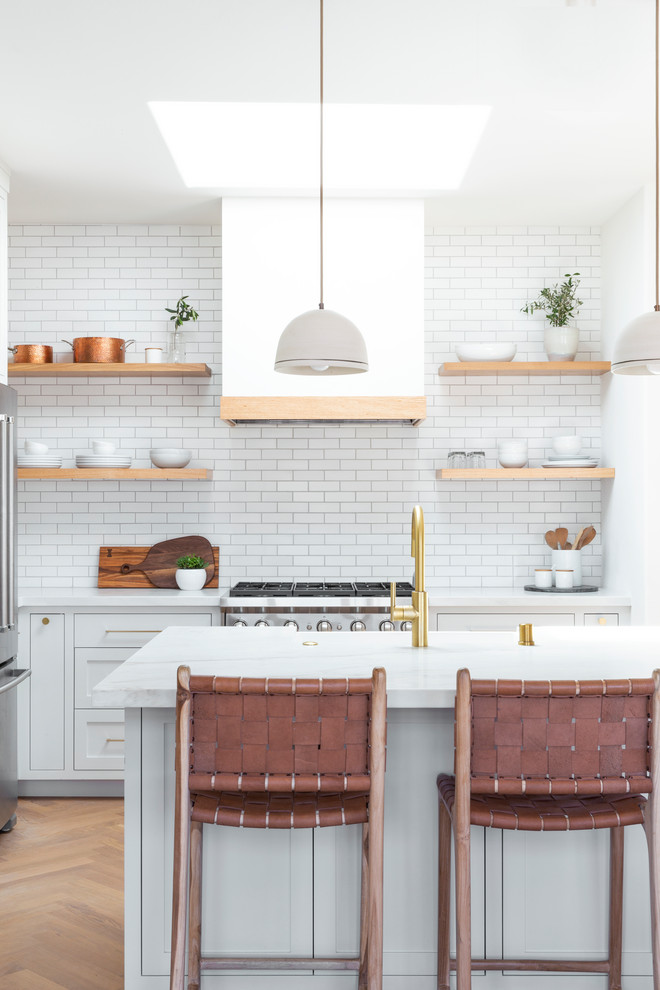 Photo of a small scandinavian l-shaped open plan kitchen in San Francisco with a farmhouse sink, shaker cabinets, white cabinets, quartzite benchtops, white splashback, subway tile splashback, stainless steel appliances, light hardwood floors, with island, beige floor and white benchtop.